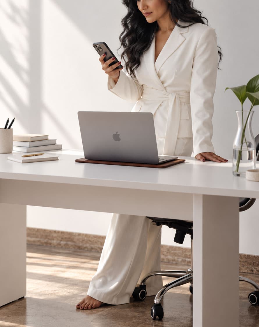 Woman reading her phone at a white desk with a laptop